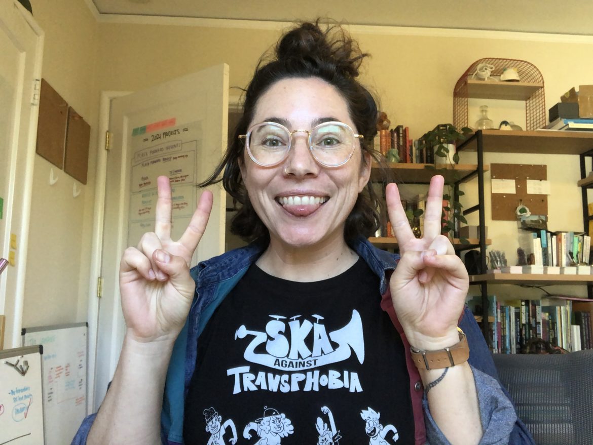 a white person wearing round glasses looking at the camera, sticking out their tongue, and holding up two peace signs -- the background is a messy office