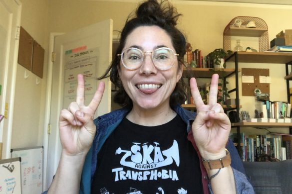 a white person wearing round glasses looking at the camera, sticking out their tongue, and holding up two peace signs -- the background is a messy office