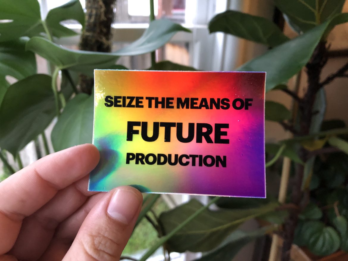 a hand holds a colorful sticket which says "Seize the means of future production" and in the background there are plants